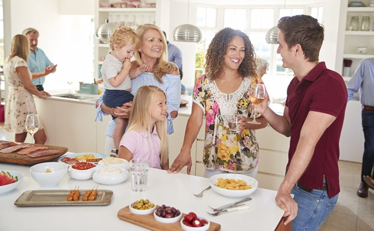 A multi-generational family is gathered in a bright kitchen, enjoying food and conversation during a social event.